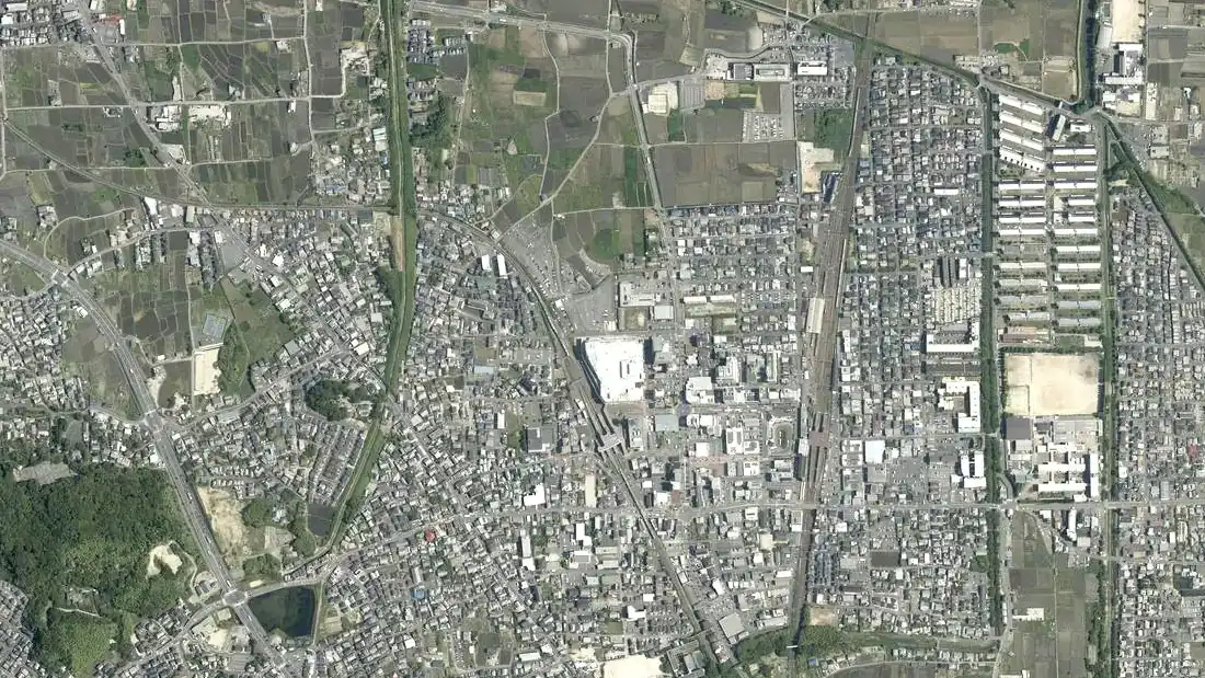 京田辺市中心部の空中写真。南北に走る2本の線路と駅前の街のまわりに、戸建ての屋根が密集する。左上に田んぼ、右に集合住宅の棟