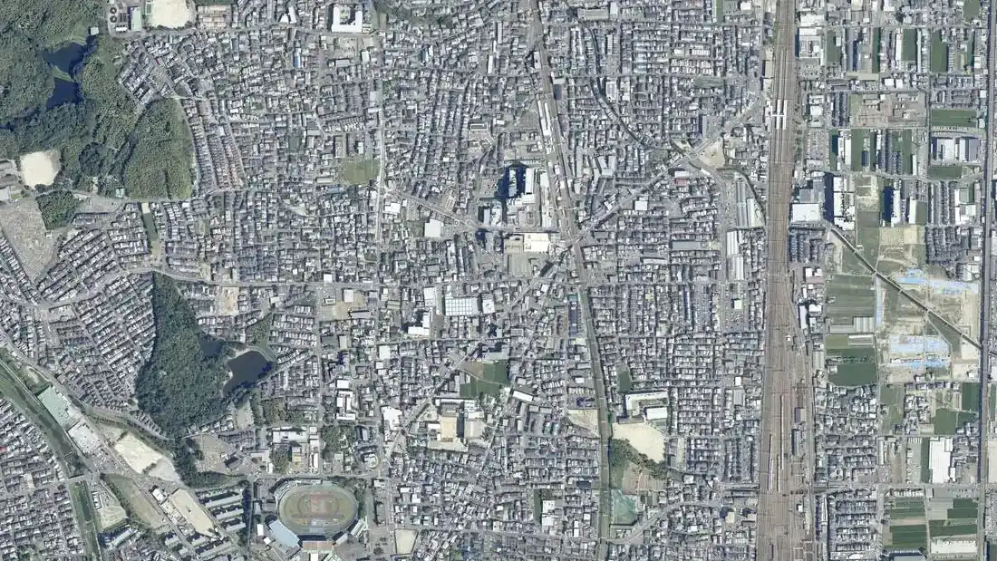 向日市中心部の空中写真。細い道に沿って戸建ての屋根がすき間なく並ぶ市街地。右に線路の敷地、左に緑地と池