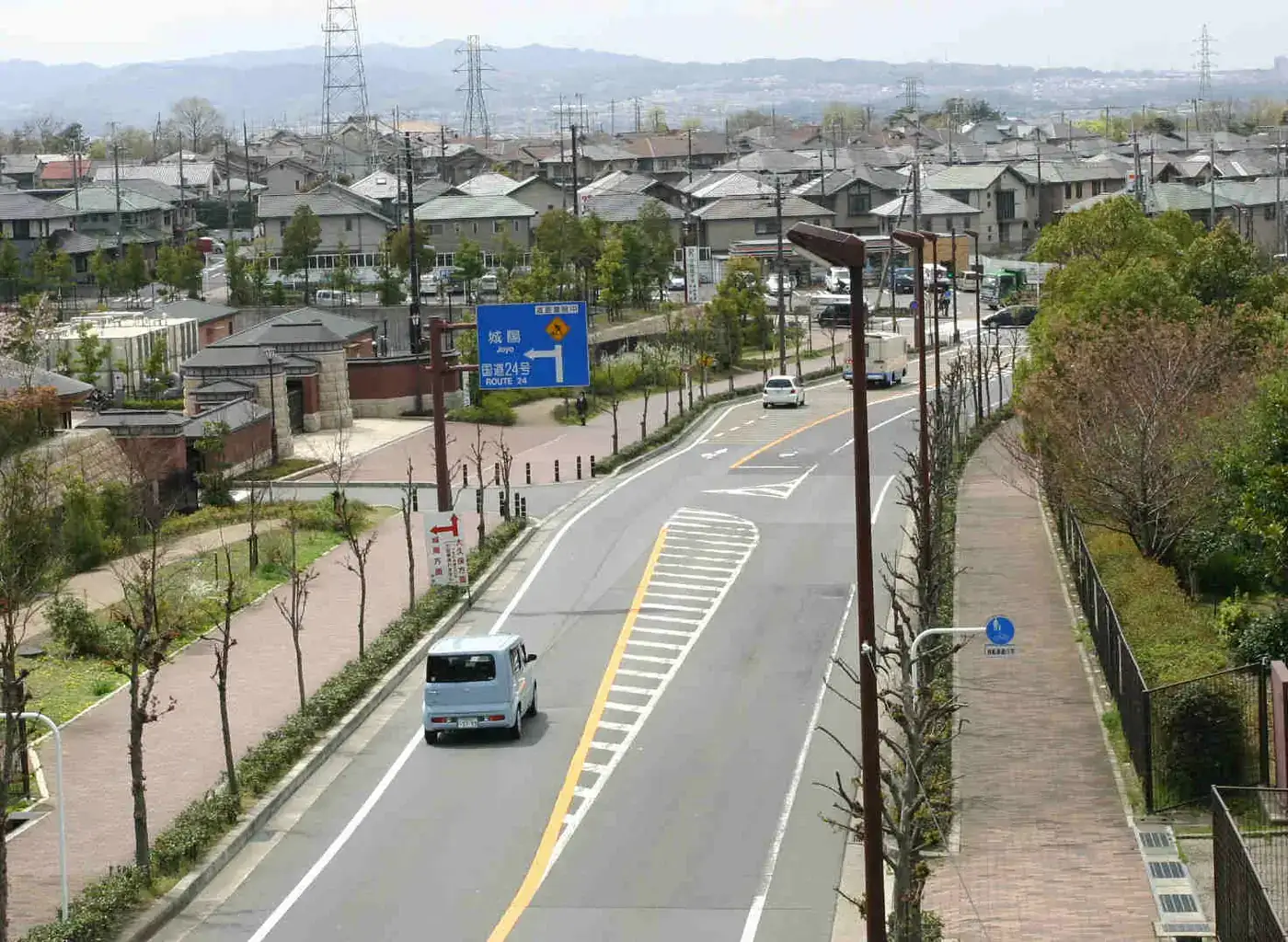 丘の上から見下ろす宇治市の住宅地。瓦屋根の住宅が並ぶ市街地と、手前に片側2車線の道路、案内標識、軽自動車1台